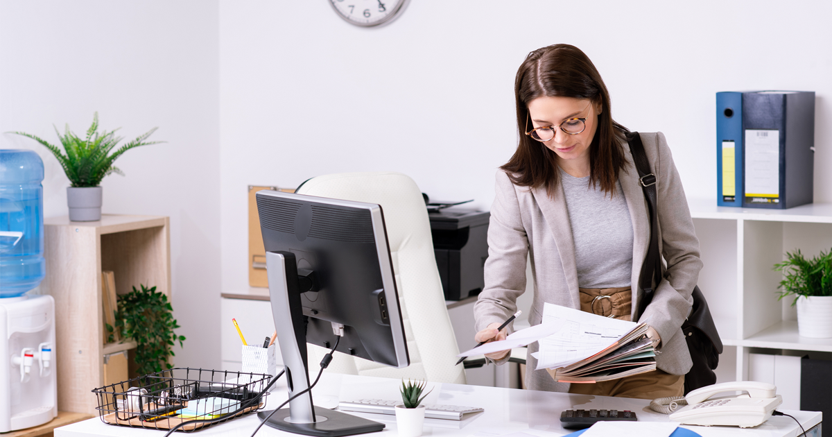 businesswoman with handbag on shoulder looking through papers while leaning over desk in office