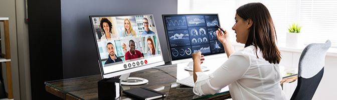 Business woman participating in a virtual conference, sitting in front of two computer screens that show attendees.