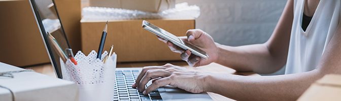 Business owner sitting at a desk in front of a laptop while looking at a mobile phone.