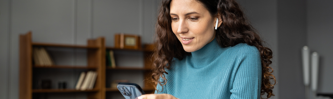 Smiling woman sitting at office desk looking at phone with earbuds in ears listening to podcast. 