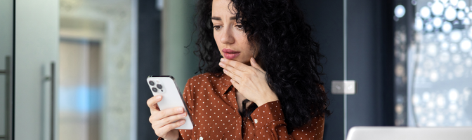 Shocked young business woman sitting in office at desk with laptop, holding phone, hand on face