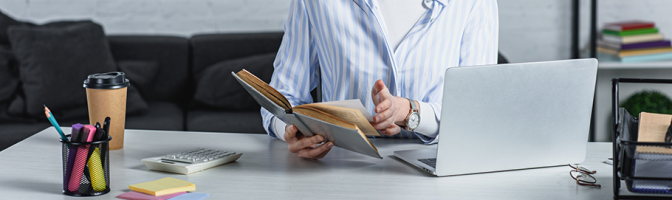 cheerful woman reading book near laptop in modern office