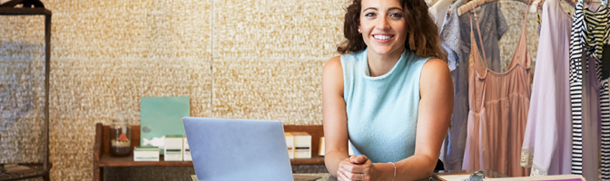 Young woman working in clothes shop leaning on counter with open laptop. 