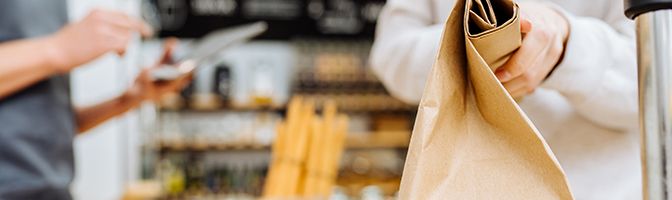 Small business employee use a tablet in the background while another employee holds a paper bag.