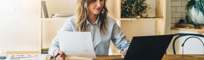 younger woman with light hair references document and laptop at desk