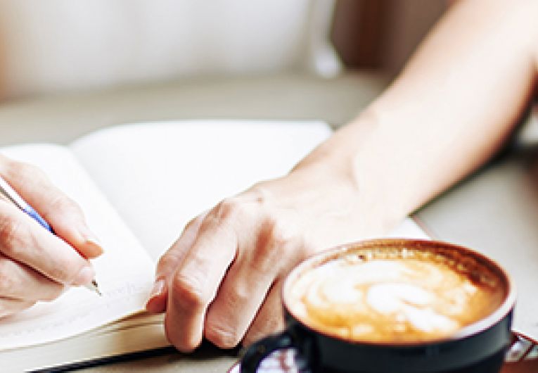 Closeup of a person writing in a journal with a cup of coffee and glasses on a table.