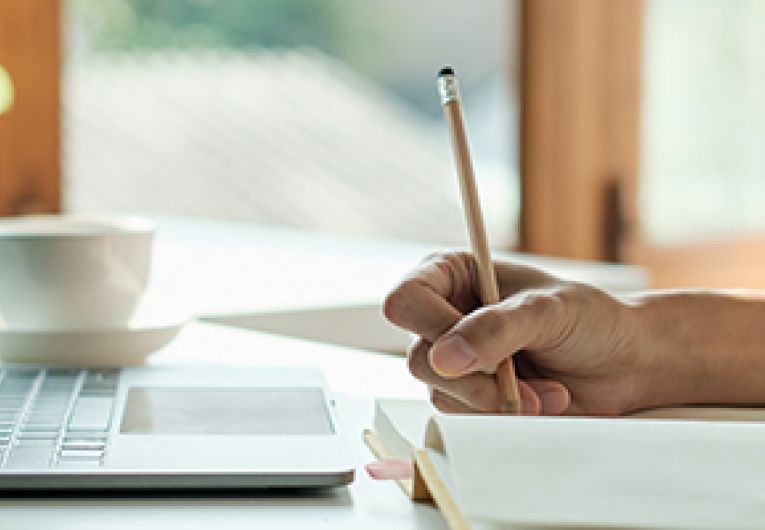 A person taking notes while viewing a laptop.