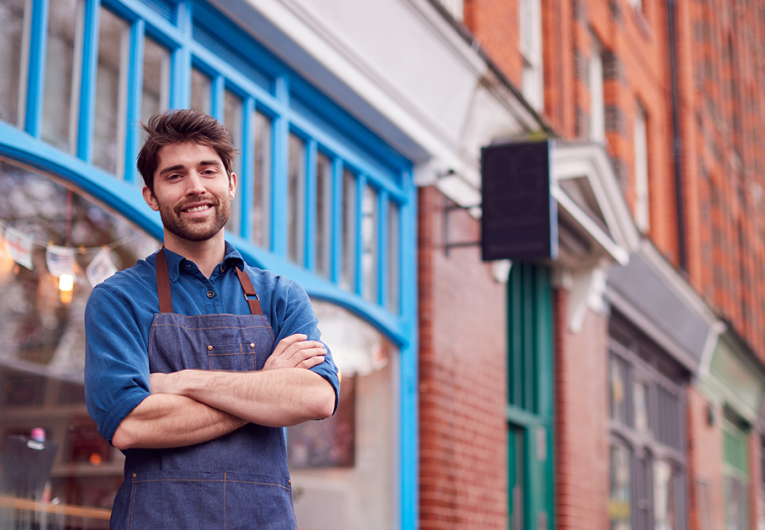 Portrait Of Male Small Business Owner Wearing Apron Standing Outside Shop On Local High Street