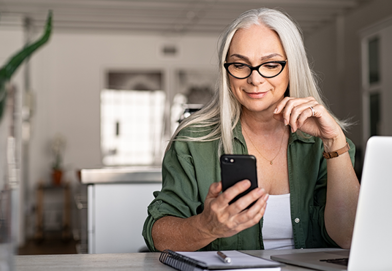 Happy senior woman using mobile phone while working at home with laptop.