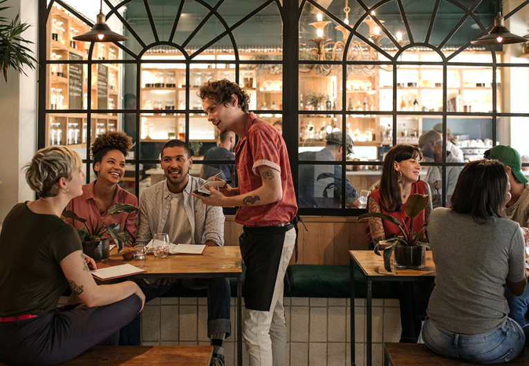 Smiling young waiter taking orders from a diverse group of customers sitting together at a restaurant table