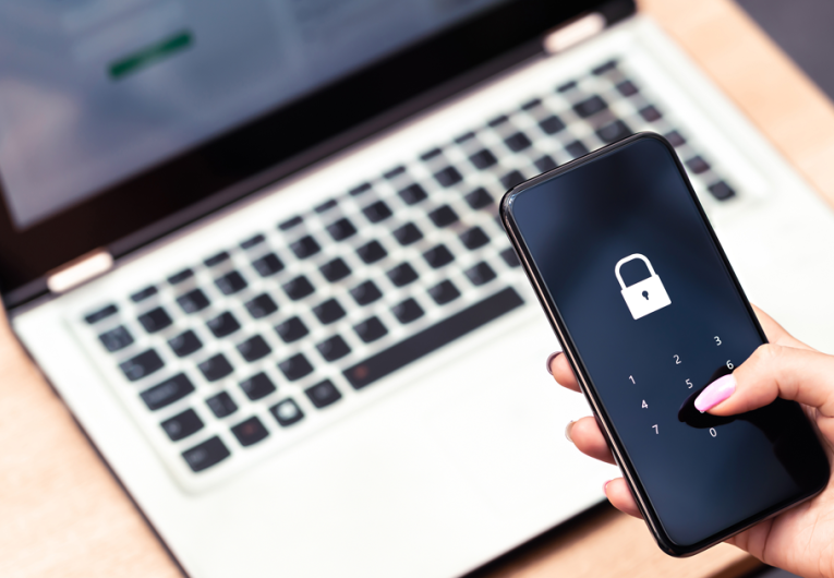 Hand holding a phone hovering over laptop computer with a password lock on the screen. 