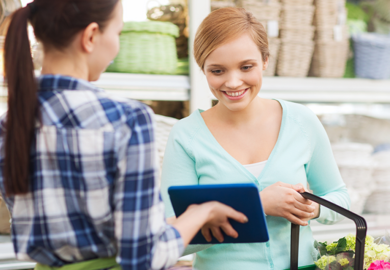happy gardener with tablet pc helping woman with choosing flowers at flower shop