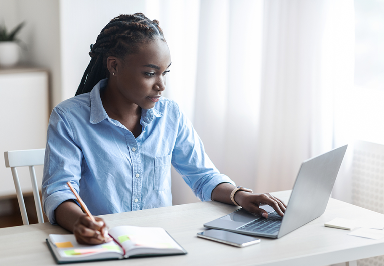 Woman Working On Laptop At Home Office And Taking Notes, Sitting At Desk Near Window, Looking At Screen, Writing Down Information From Computer