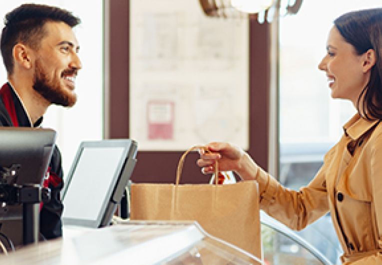 A small business owner smiles as he hands over a shopping bag to the customer at the register.