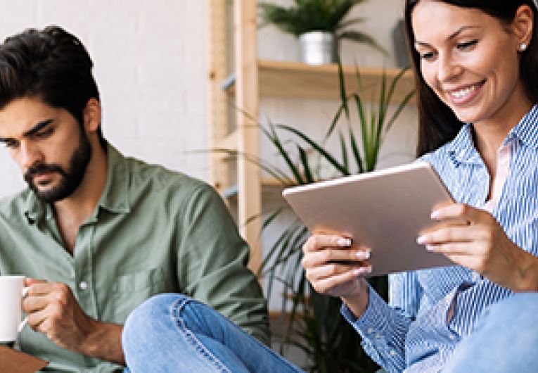 A woman reads an eBook while a man reads a book as he holds a cup in one hand.