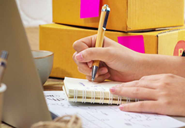 Close up of a small business owner writing in a notebook at a desk in front of a laptop.