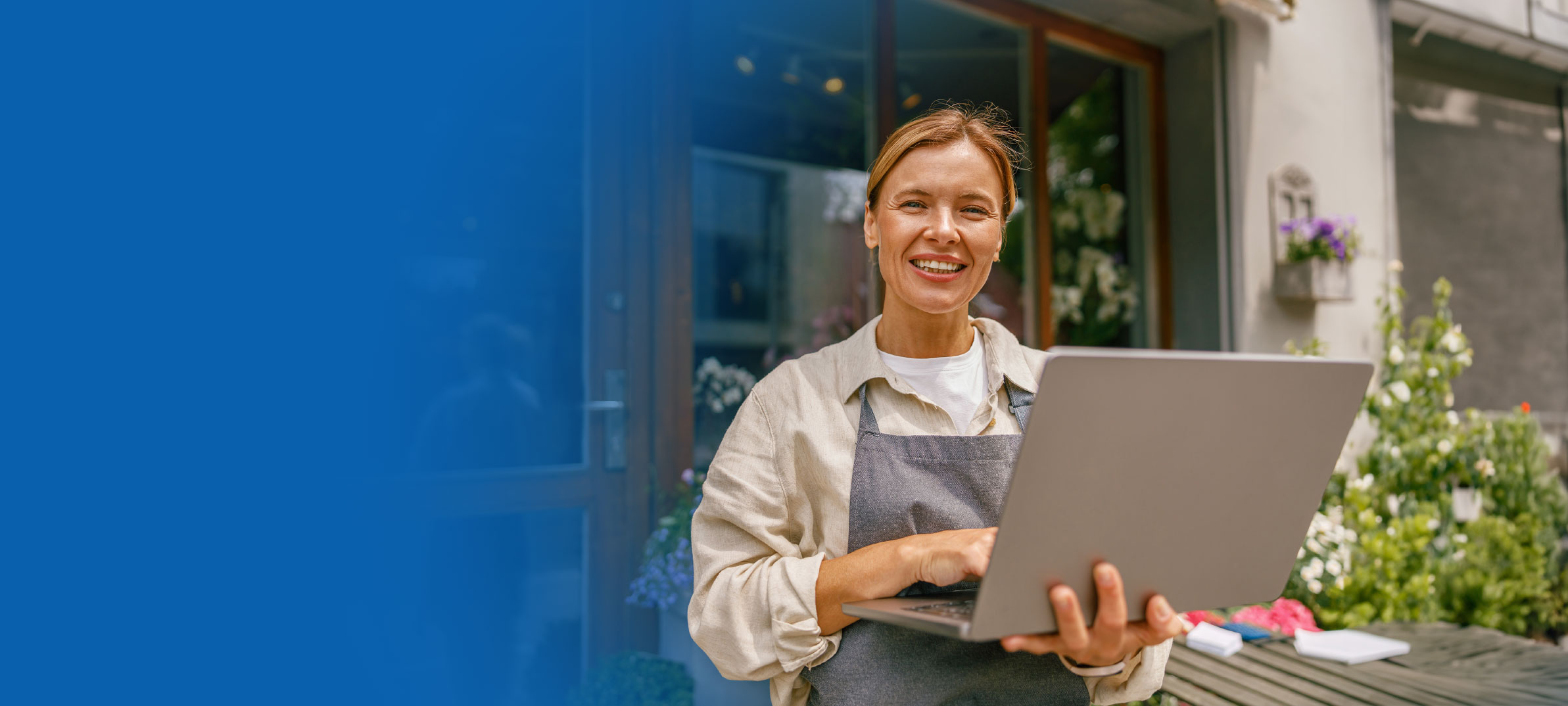 Woman wearing a work apron smiling and holding a tablet outside a business