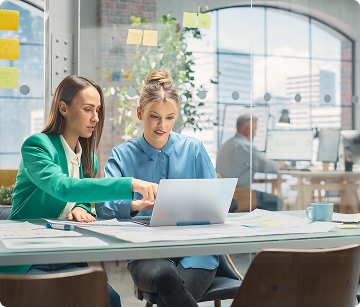Two women sitting at a table looking at a laptop in a large office